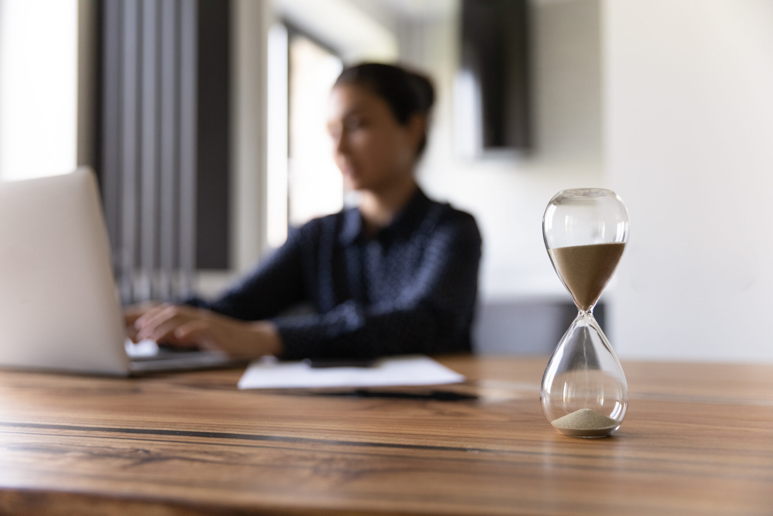 An hourglass in focus on a desk while a woman types in the background—symbolizing the power of short, focused time blocks to spark creativity.