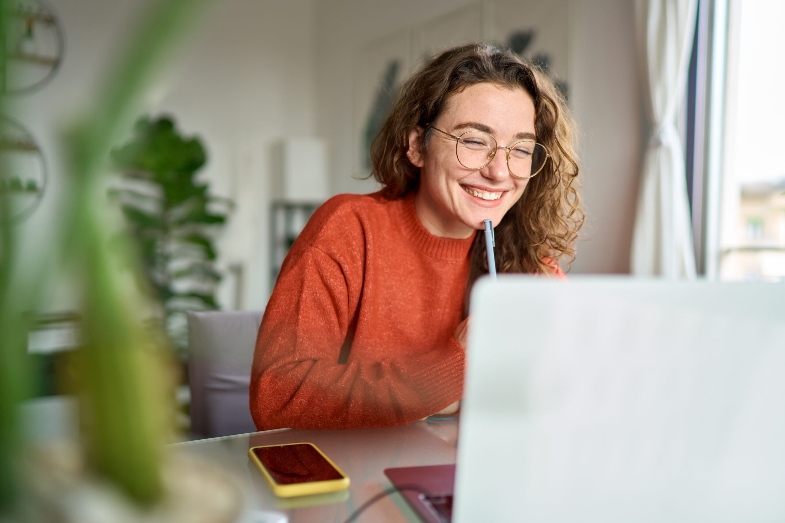 A cheerful woman smiling at her laptop with a pen in hand—reflecting the joy and growth that comes from consistently practicing creativity.