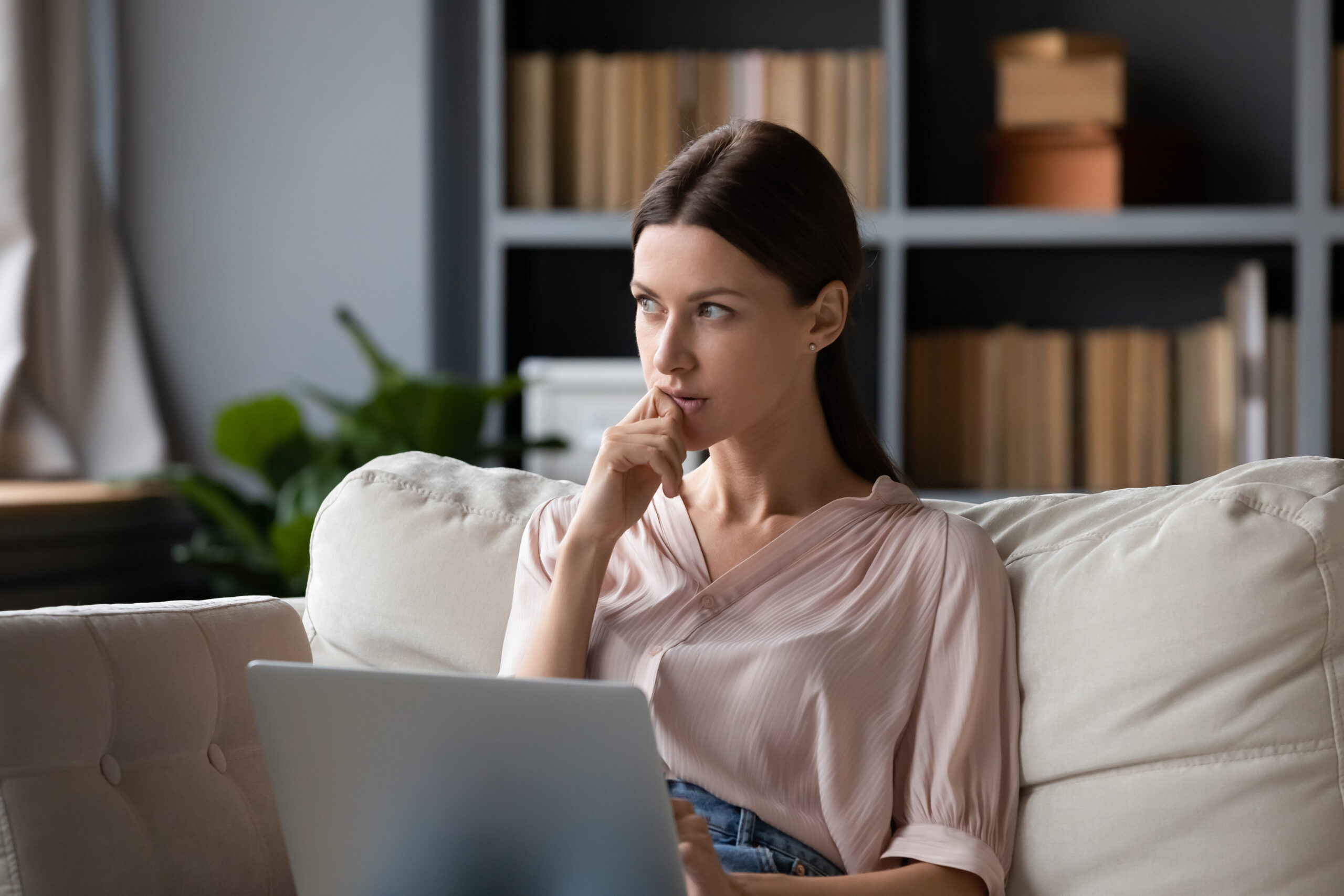 A thoughtful woman sitting on a couch with a laptop, lost in reflection—capturing the moment before rediscovering the joy and freedom of playful creativity.